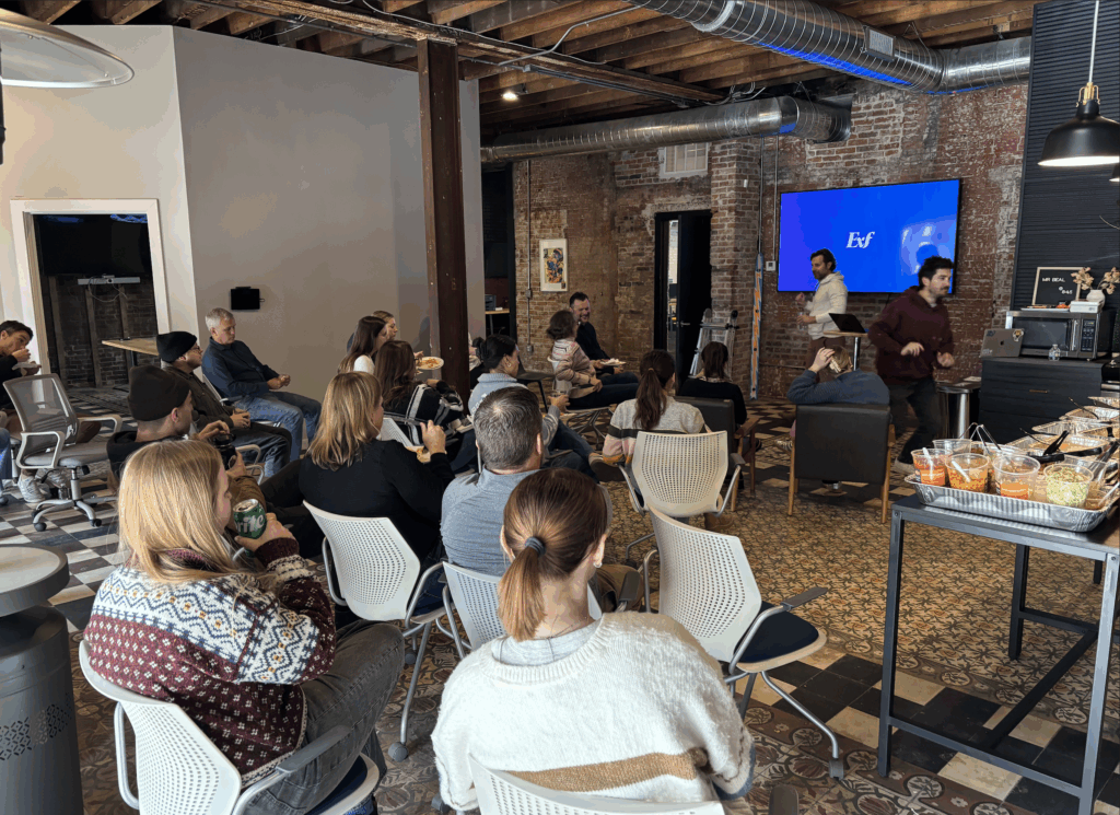 A group of people sit in an office space, facing a screen with EoF displayed. Two people stand at the front, with snacks on a table nearby. The room has exposed brick walls and ceiling beams.