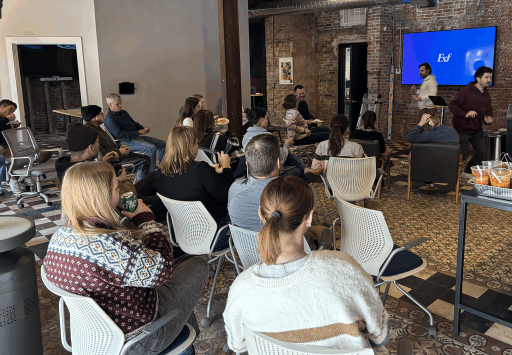 A group of people sit in an office space, facing a screen with EoF displayed. Two people stand at the front, with snacks on a table nearby. The room has exposed brick walls and ceiling beams.