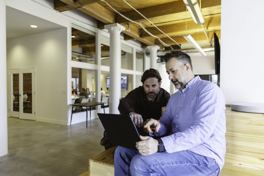 Two men sitting indoors on wooden steps, collaborating and looking at a laptop. One points at the screen while the other listens attentively. Office environment with exposed beams and glass-walled meeting rooms in the background.