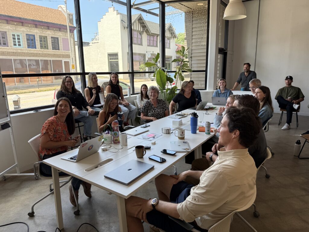 A group of people sit and smile around white tables in a modern, bright office space with large windows, laptops, notebooks, and coffee cups, appearing engaged in a casual meeting or discussion.
