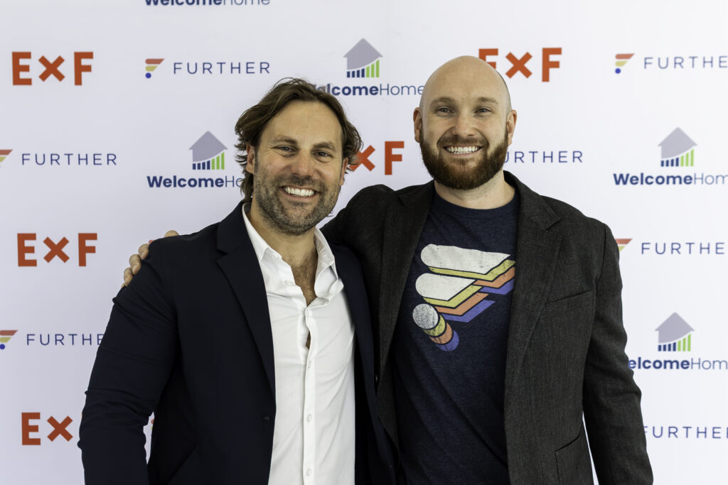 Two smiling men stand together in front of a step-and-repeat banner with logos for ExF, Further, and WelcomeHome. One man wears a suit and the other wears a blazer over a graphic T-shirt.